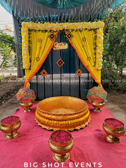 A complete outdoor Haldi ceremony setup featuring a vibrant yellow drape backdrop, a custom neon "Haldi" sign, and traditional Rajasthani-style umbrellas. The central seating area includes a golden lotus-shaped basin and brass bowls filled with flower petals, all set on a bright pink carpet for a colorful and festive pre-wedding celebration.