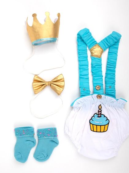 A baby boy's first birthday outfit, featuring a cupcake-themed romper with suspenders, socks, a bow tie, and a crown.