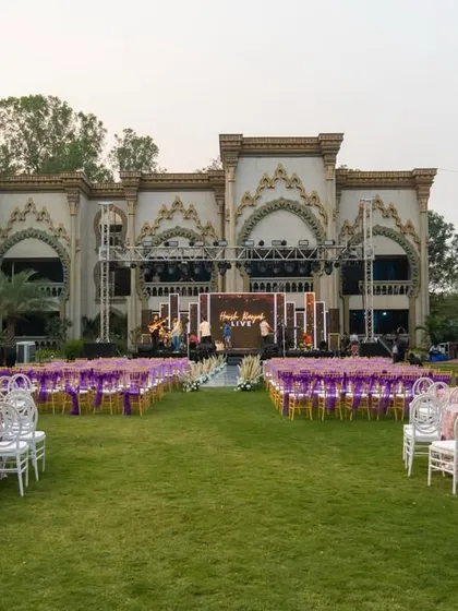 For a grand 25th-anniversary celebration, I transformed this lawn into an elegant affair. The setup includes a large stage, beautiful seating arrangements with purple and white themes, and a stunning palace-like venue in the background.