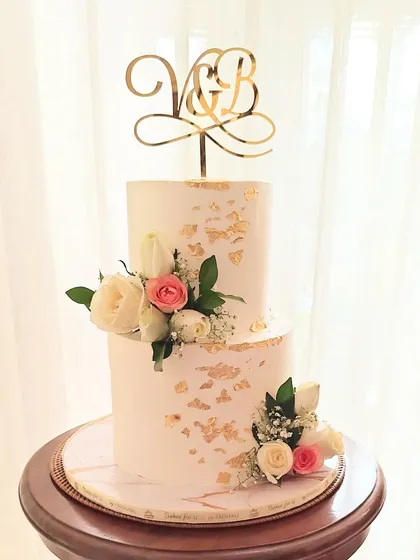 An elegant two-tier engagement or wedding cake with gold leaf accents and arrangements of fresh pink and white roses. The custom initial topper adds a personal touch.