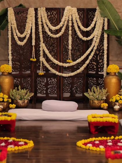 A traditional Mehendi setup with a carved wooden screen, jasmine garlands, and banana trees. The low seating on the floor creates a classic, intimate atmosphere for the function.