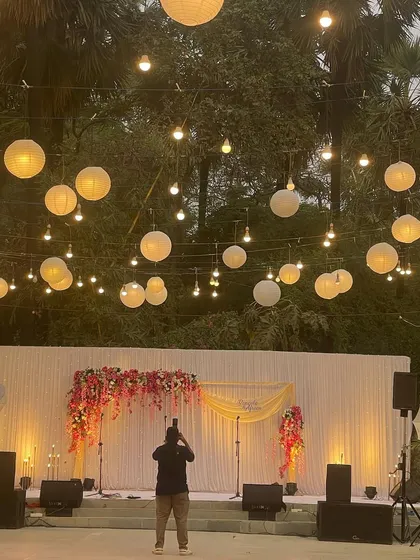 A view of a reception stage from afar, with a photographer capturing the moment. The canopy of hanging lanterns and string lights creates a magical, starlit sky effect.