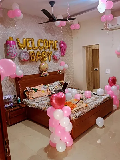 A cozy bedroom setup to welcome a new baby. The wall is decorated with "WELCOME BABY" foil balloons, along with pink and white balloons and baby-themed foil shapes like a bottle and footprint.