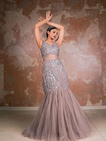 A full-length shot of a model in a grey mermaid-style gown. The pose is designed to accentuate the dress's silhouette and the intricate silver embroidery, set against a textured backdrop for a touch of artistic drama.