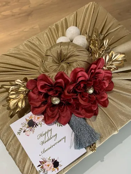 The final touch is everything. Here, an anniversary gift is elegantly wrapped in golden satin fabric using the Furoshiki technique, adorned with rich red flowers and a tassel for a luxurious finish.