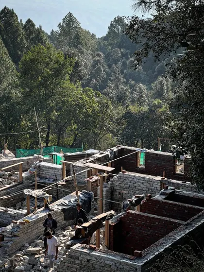 An aerial view of the construction site in the mountains, showing the foundations of several guesthouses being laid out amidst piles of locally quarried stone. The project is emerging from the earth itself.