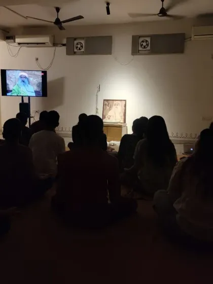A gathering for a joint Sadhguru Sanidhi Process and Devi Abhishekam. The room is dark, with all focus on the Guru's presence on screen and at the altar.
