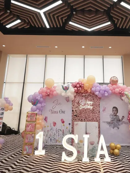 A grand butterfly-themed setup for Sia's first birthday. The decor features a large balloon arch in pastel shades, a sequin wall, and personalized backdrops against a modern chevron-patterned floor.