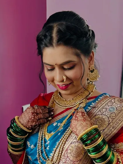 A candid moment of the bride adjusting her beautiful gold jewellery. Her makeup features soft pink tones on the eyes and lips, complementing her red and blue saree.