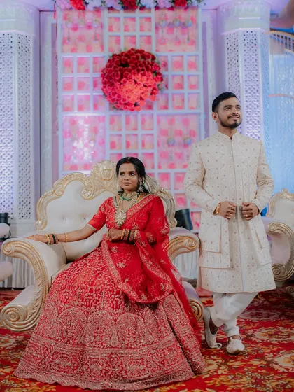 A regal portrait of the couple at their reception. The bride is seated on a throne-like chair while the groom stands beside her, creating a classic and elegant wedding portrait.