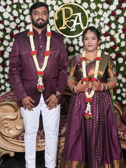 A formal portrait of an engaged couple against a floral wall. The bride's makeup is sophisticated, matching her deep purple langa davani and the overall elegant theme.