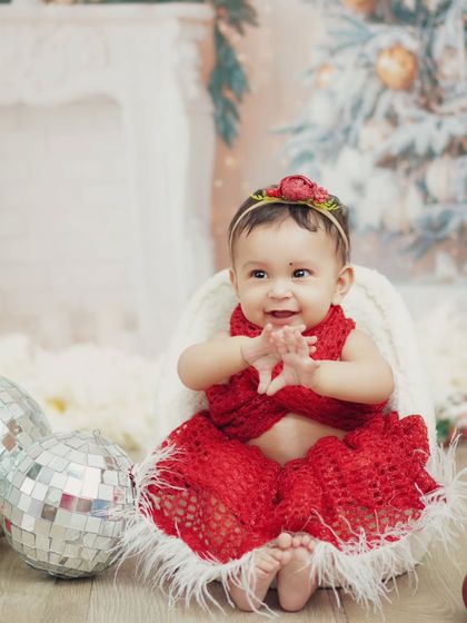 A beautiful baby girl in a festive red outfit for her first Christmas photoshoot. The soft, snowy backdrop creates a magical winter wonderland feel.