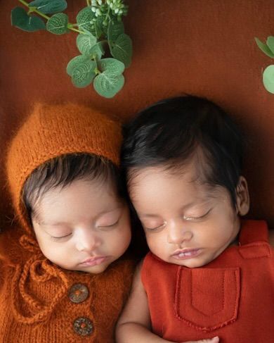 A close-up of newborn twins sleeping peacefully side-by-side. The warm, earthy tones of their outfits and the natural greenery create a serene and timeless portrait.