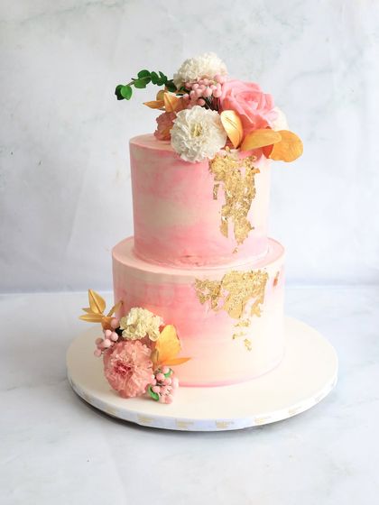 A two-tiered cake with a soft pink watercolor finish and touches of edible gold leaf. The design is completed with delicate arrangements of fresh and sugar flowers for a romantic and artistic feel.