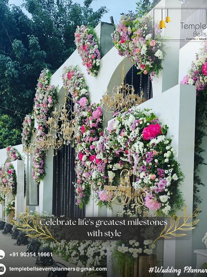 We transform venues into masterpieces. This decor features a white facade adorned with cascading pink florals and ornate golden chandeliers for a luxurious feel.