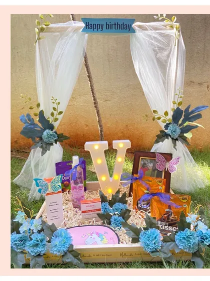A grand initial floral hamper for a birthday celebration. This gift features a large light-up letter 'V', a beautiful canopy-style decoration, and is filled with blue-themed treats and flowers.