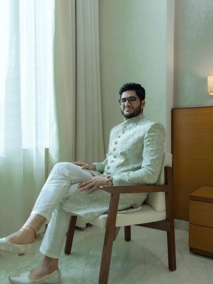 A full-length portrait of the groom seated, looking calm and ready. His light green sherwani and relaxed posture create a picture of modern elegance.