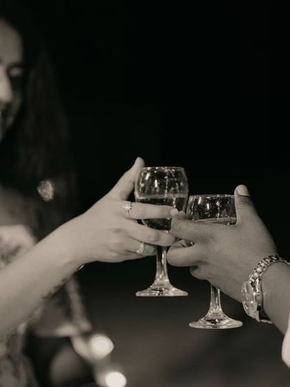 A close-up, black and white shot of a couple toasting with wine glasses during their romantic beach picnic, focusing on the detail of their hands and the engagement ring.