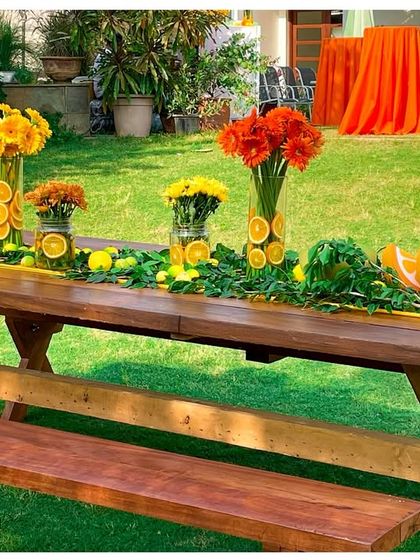 A rustic wooden picnic table decorated with a long, lush centrepiece of flowers, greenery, and fruit props. This setup is perfect for an outdoor garden party.