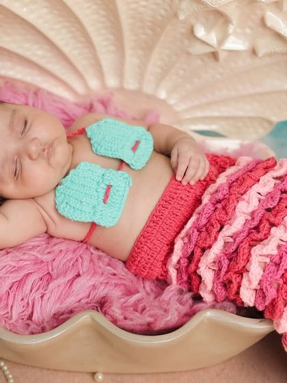 A magical 'Baby Mermaid' theme. This newborn is dressed in a crocheted mermaid tail, sleeping peacefully in a giant seashell prop against a serene ocean backdrop for a truly enchanting portrait.