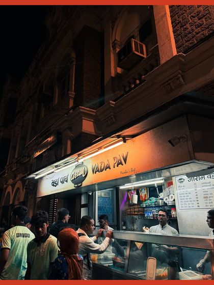 The warm light of a Vada Pav stall spills onto the street, illuminating the crowd against the backdrop of a grand old building.