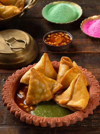 A Holi-themed shot of samosas served in a terracotta dish with chutneys. The background styling with colored powders adds a festive, celebratory context.