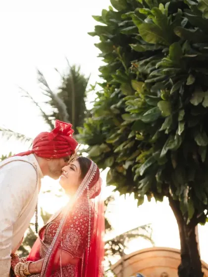 A classic wedding portrait with a sun-kissed flare. The bride in her traditional red lehenga shares a kiss with her groom, a timeless image of love and commitment.