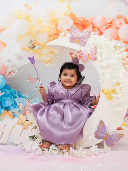 A magical first birthday portrait of a little girl in a purple dress, holding a wand on a butterfly-adorned moon.