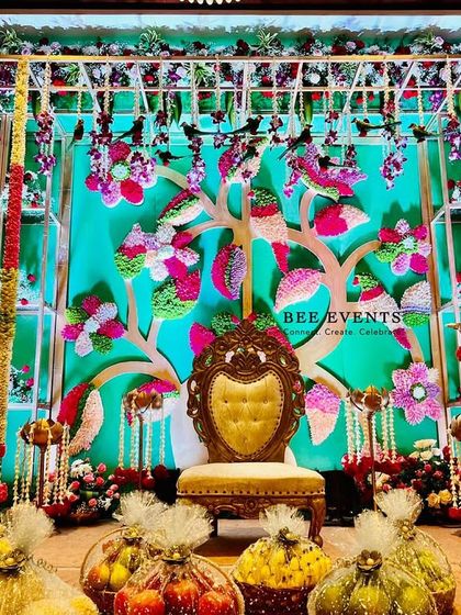 A vibrant and colorful stage setup for an engagement or pre-wedding ceremony. The backdrop features a 'tree of life' motif adorned with multi-colored flowers, surrounded by traditional elements and fruit baskets.