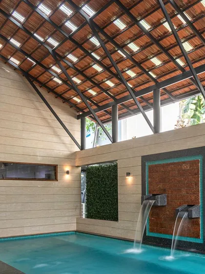 This indoor swimming pool features two waterfalls cascading down a textured mosaic wall. The space is covered by a traditional tiled roof with skylights, creating a private, tropical oasis feel.