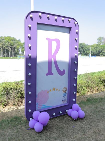 A light-up marquee sign with the initial 'R' for a personalized touch at an outdoor carnival party. The purple frame and small balloon cluster add a pop of color.