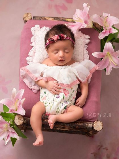 A sweet baby girl on a tiny wooden bed, surrounded by pink lilies. The floral elements add a touch of natural beauty to the scene.