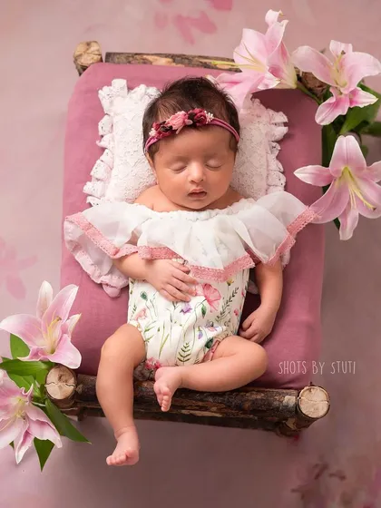 A sweet baby girl on a tiny wooden bed, surrounded by pink lilies. The floral elements add a touch of natural beauty to the scene.