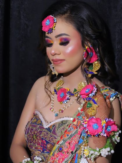 A close-up of a vibrant Haldi or Mehendi look. The colorful eye makeup is perfectly complemented by the bright pink floral jewellery, including the maang tikka and earrings.