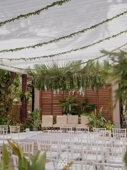 A clean and simple mandap design for a minimalist aesthetic. The setup uses a canopy of white fabric with green garlands, white seating, and a backdrop of natural wood and plants.