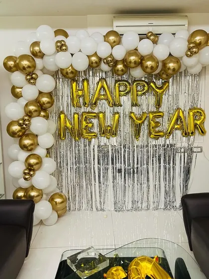 A classic "Happy New Year" decoration for a home or office party. The setup includes a silver tinsel curtain and a white and gold balloon arch.