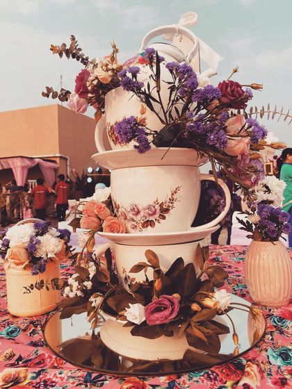A creative centerpiece for an 'Alice in Wonderland' tea party. A stack of oversized, floral-patterned teacups overflows with beautiful purple and pink flowers, perfectly capturing the theme's playful spirit.