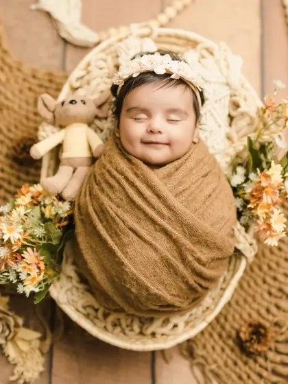 A smiling newborn in a brown swaddle, holding a tiny teddy bear in a boho-style setup.