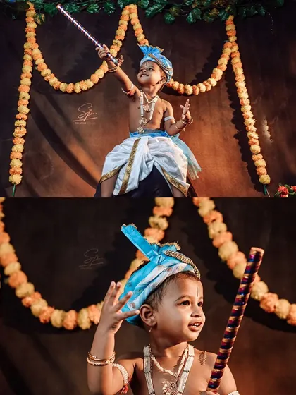 A full-body shot showing the complete little Krishna costume and pose. The marigold garland backdrop and traditional props create an authentic festive atmosphere for this creative kids' portrait.