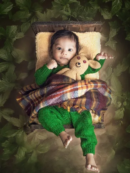 A two-month-old baby, wide-eyed and curious, snuggled in a tiny bed with his teddy bear. The green knit outfit and leafy surroundings give this a cozy, natural feel.