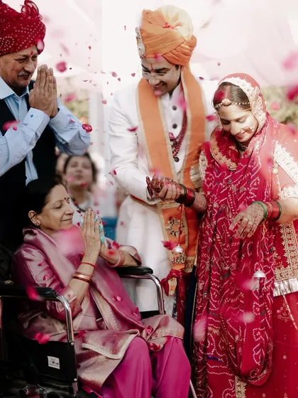 During a traditional Indian wedding ceremony, a couple receives blessings from an elder family member in a wheelchair, as rose petals shower down around them. This is a powerful, emotional moment of intergenerational love and respect.