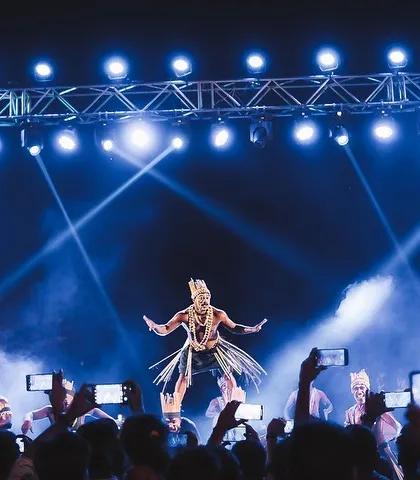 A wide shot shows a tribal dancer in mid-air, surrounded by an audience capturing the moment on their phones. This image encapsulates the entire atmosphere of a modern cultural event: the stunning performance, the dramatic lighting, and the engaged crowd.