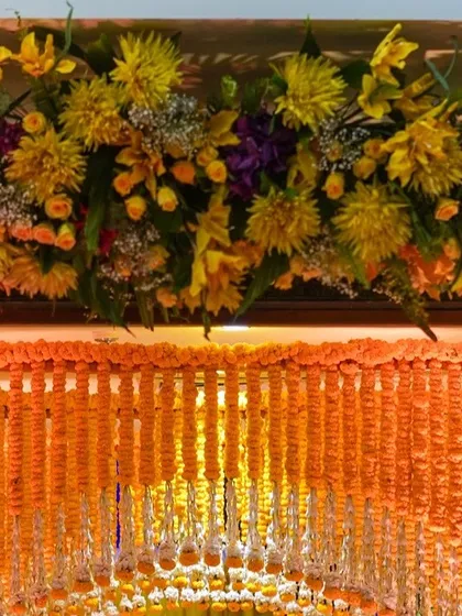 A close-up of the top of a floral arch, showing a mix of yellow flowers and baby's breath. Below, a dense curtain of marigold strings creates a classic Haldi entrance.