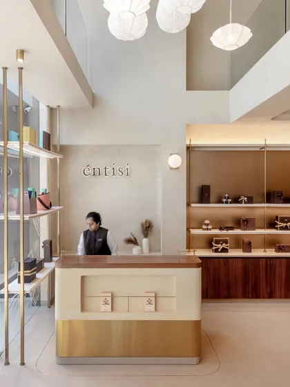 The reception counter at Entisi combines a terrazzo-finish body with a warm wood top and a brushed brass base, establishing the store's material language right at the entrance. The double-height ceiling and minimalist pendant lights create a sense of scale and elegance.