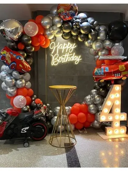 A Cars-themed 4th birthday party. This setup includes a circular balloon arch in red and silver, a "Happy Birthday" neon sign, and a toy motorbike.
