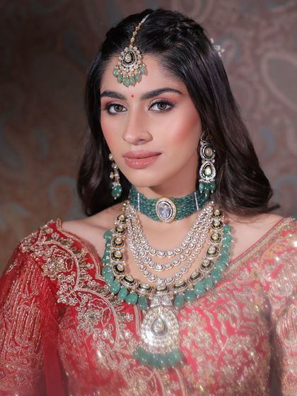 A close-up on a classic red bridal lehenga look. The makeup features a soft cut-crease on the eyes and a warm, nude lipstick, allowing the heavy emerald jewellery to shine.