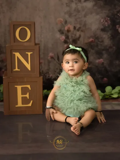 A baby girl in a cute green ruffled outfit sits next to "ONE" blocks for her first birthday milestone photos.