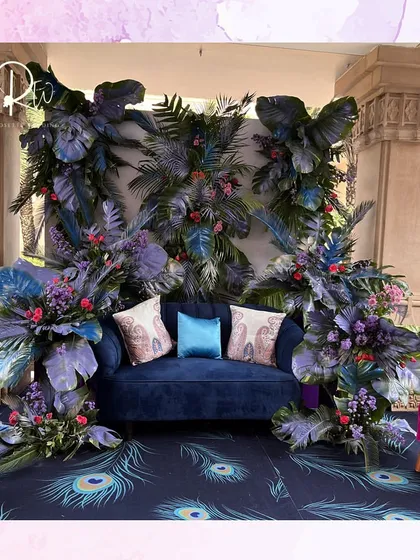 A photo booth for the peacock-themed event, with a deep blue sofa set against a backdrop of dark, tropical leaves and pops of colorful flowers.