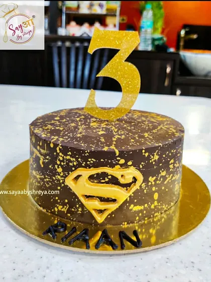 A super cake for a super third birthday. This design features the iconic Superman logo against a dark chocolate cake with a gold splatter effect, creating a simple yet powerful statement.