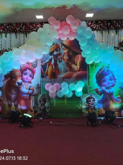 A Krishna-themed stage decoration for a naming ceremony. The backdrop features images of Krishna and a pastel-colored balloon arch.